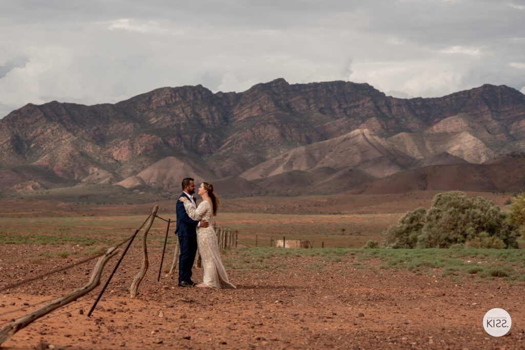 A wedding couple embracing on a red dirt track beneath mountain ranges