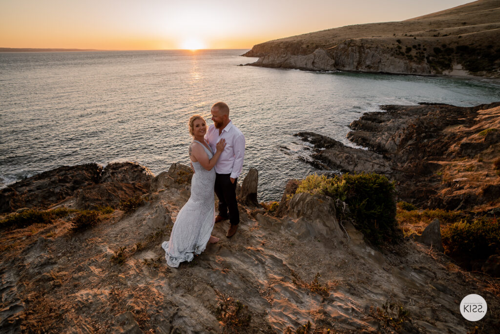A wedding couple embracing on coastal rocks at sunset