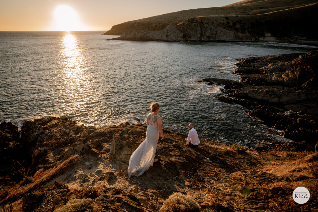 A bride standing beside her seated groom on coastal rocks