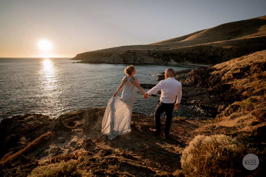 A couple walking hand in hand over coastal rocks at sunset