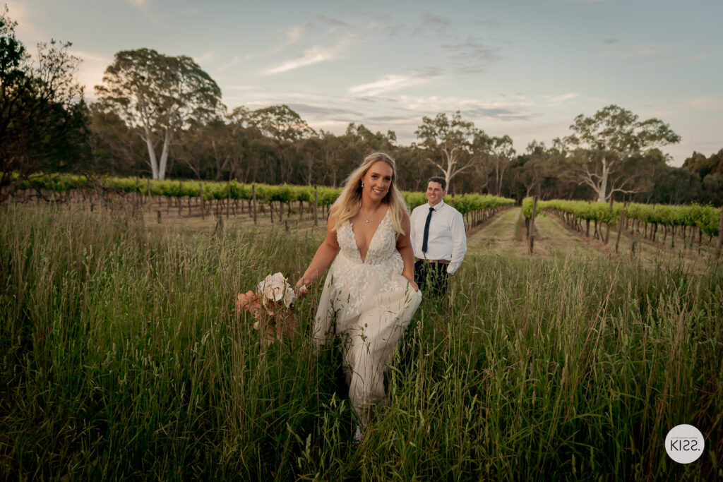 A wedding couple walking through tall grass beside vineyard rows
