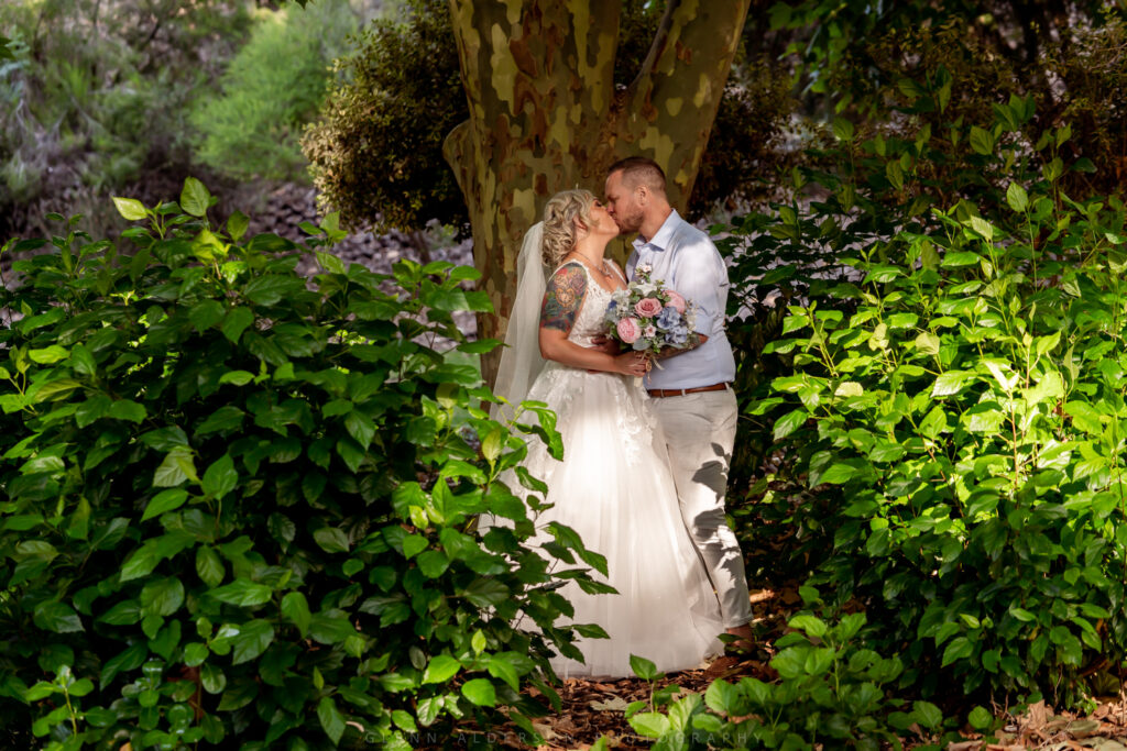 A wedding couple kissing beside a tree in a lush garden