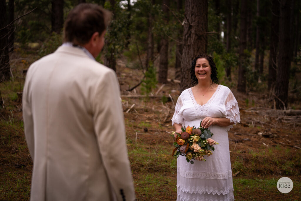 A bride smiling towards her groom in a pine forest