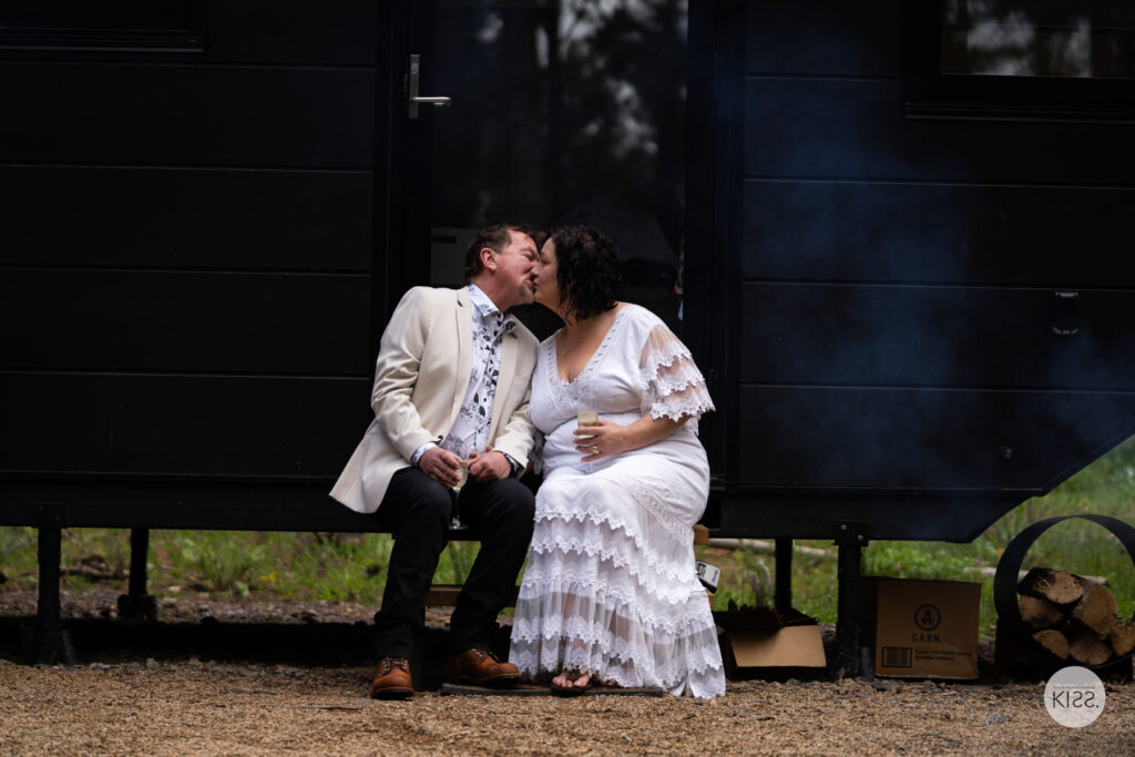 A couple kissing on a bench outside their forest cabin