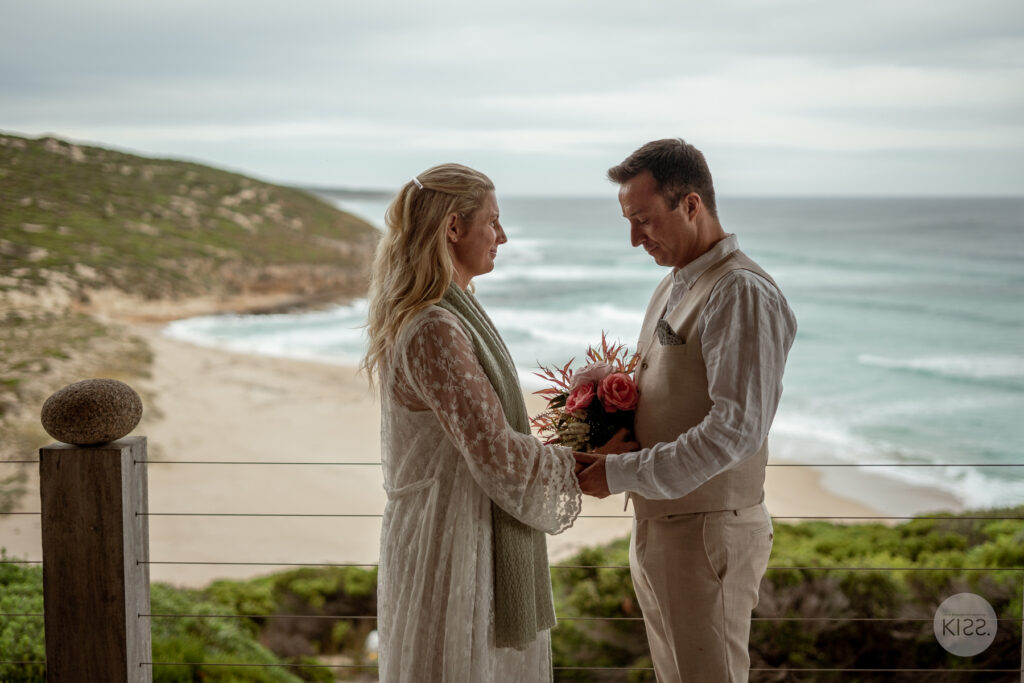 A wedding couple holding hands and flowers on a coastal deck