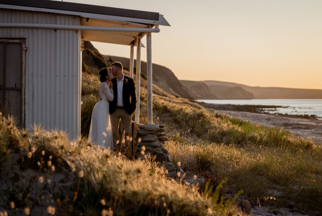 A wedding couple beside a small coastal building at sunset