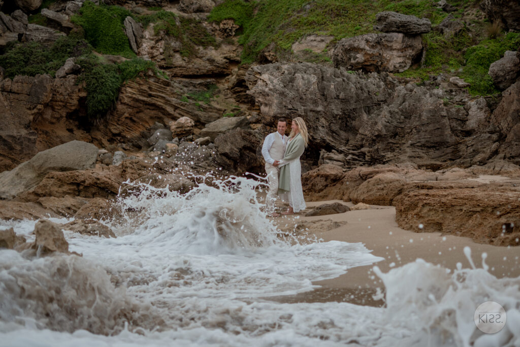 A wedding couple beside coastal rocks as a wave breaks