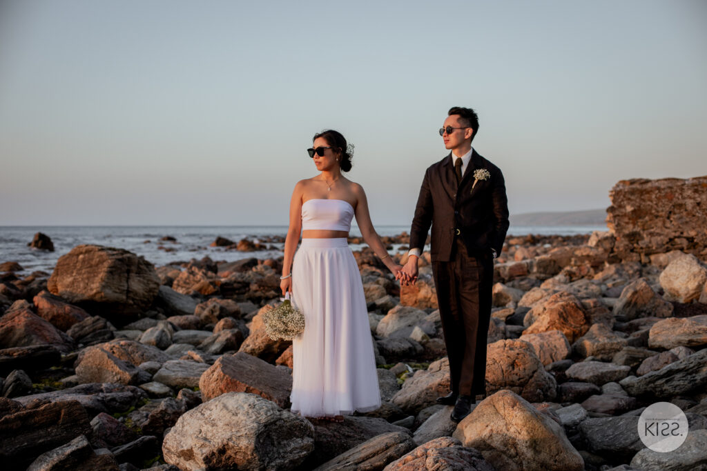 A wedding couple holding hands beside coastal rocks