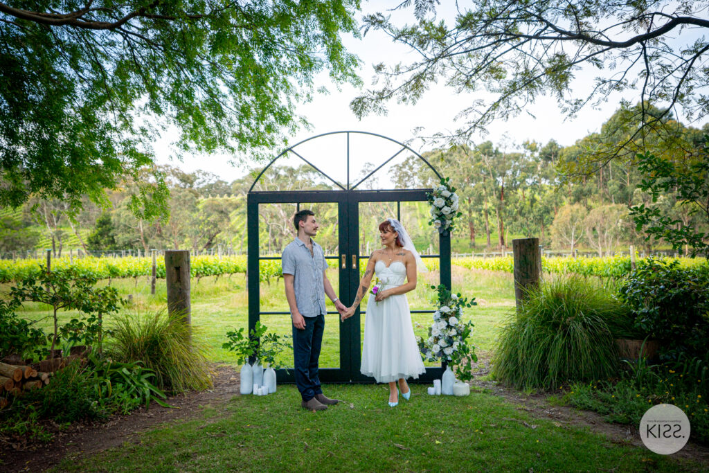 A wedding couple standing beneath a floral arbour in a vineyard setting