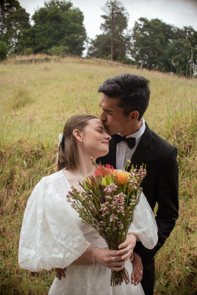 A groom kissing his bride's forehead in a grassy field
