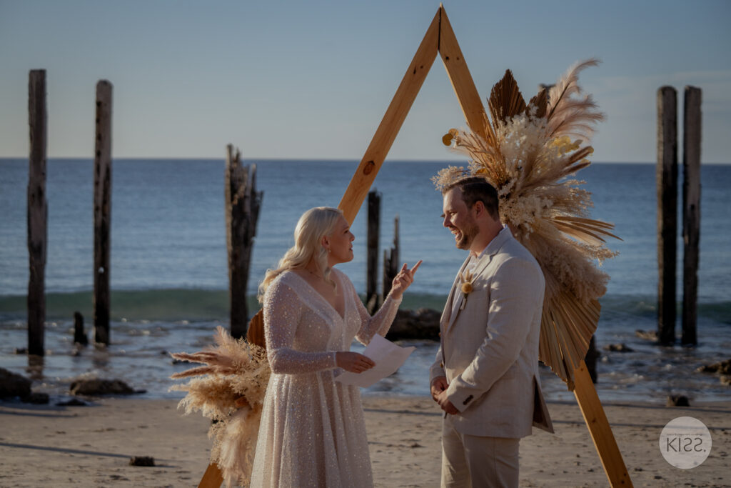 A couple exchanging vows beneath a triangular beach wedding arbour