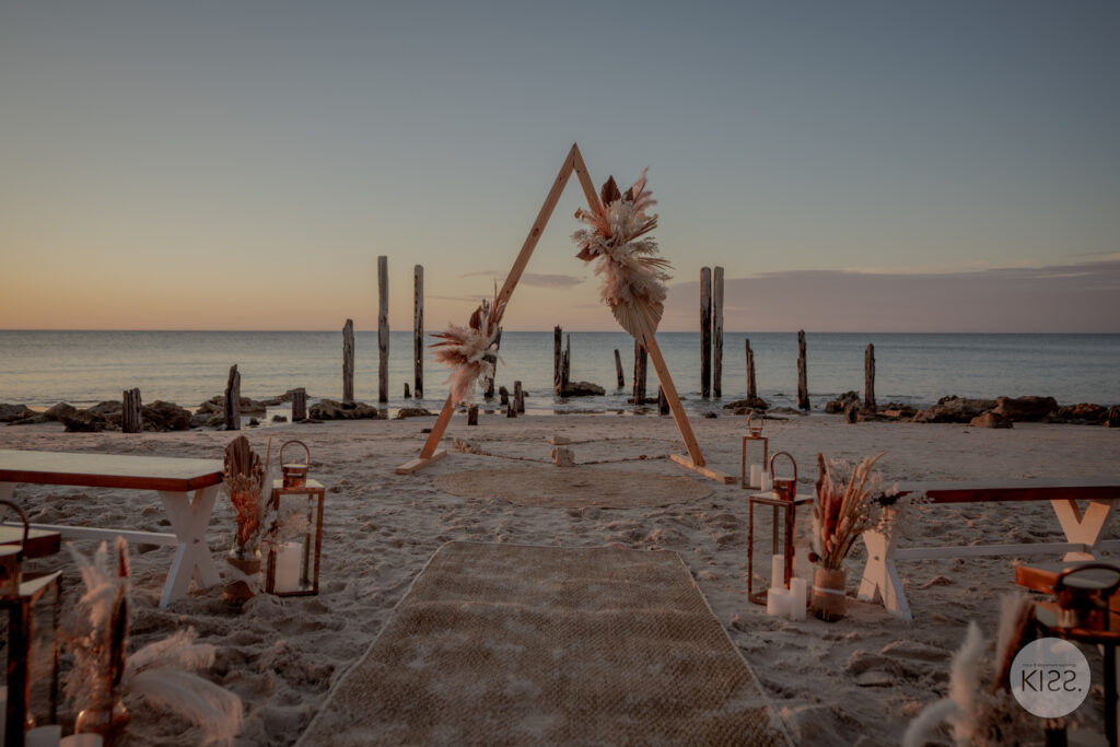 Beach wedding aisle leading to a triangular floral arbour at sunset
