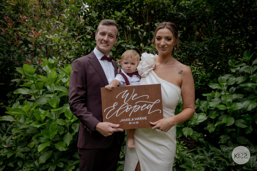 A wedding couple holding their baby and a We Eloped sign