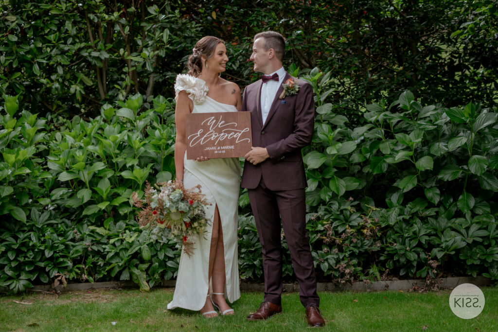A wedding couple holding an elopement sign in a garden