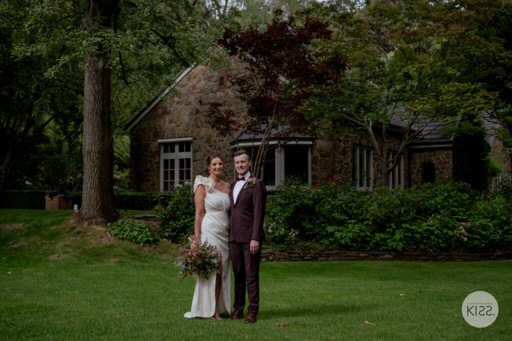 A wedding couple standing on the lawn outside a stone cottage