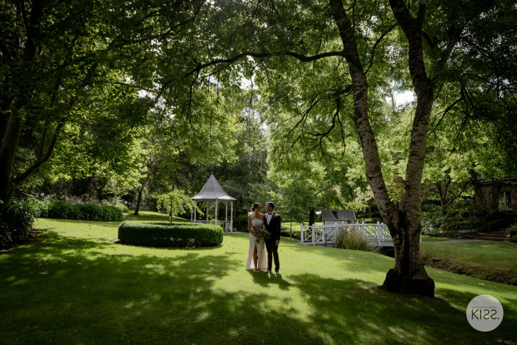 A wedding couple in a garden with mature trees and a gazebo