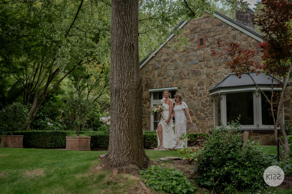 A bride standing outside a stone cottage beneath mature trees