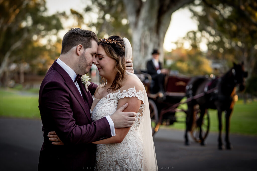 A wedding couple touching foreheads with a horse-drawn carriage behind them