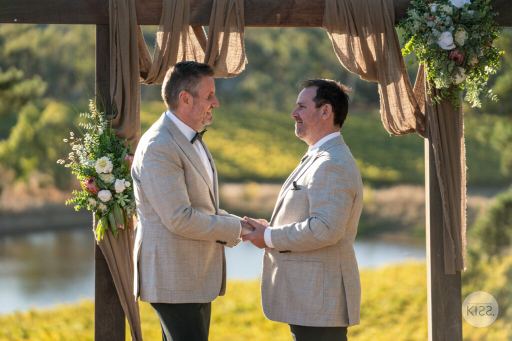 Two grooms holding hands beneath a floral arbour