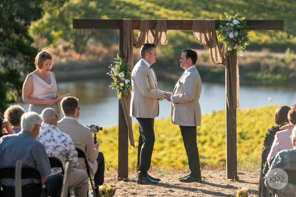 Two grooms and a celebrant beneath an outdoor wedding arbour