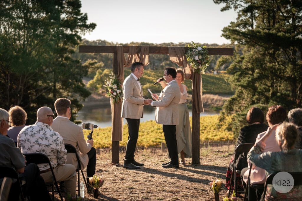 Guests watching two grooms beneath an outdoor wedding arbour