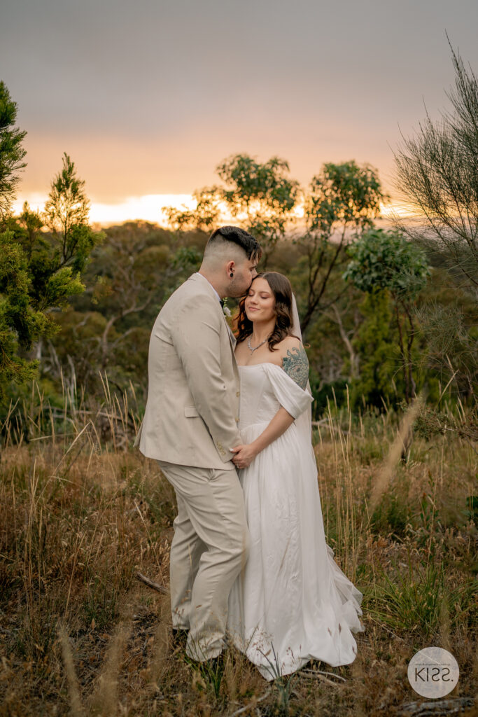 A groom kissing his bride's forehead among trees