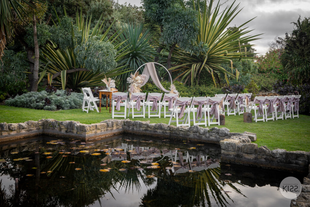 White wedding chairs and a circular arbour beside a garden pond