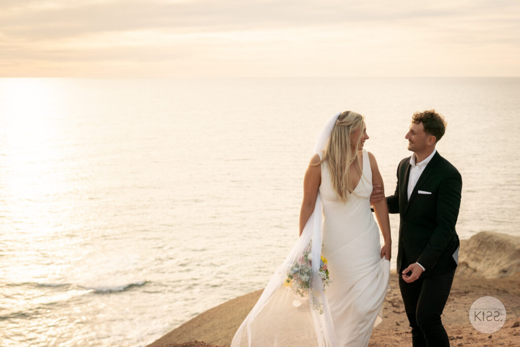 A wedding couple smiling at each other beside the sea