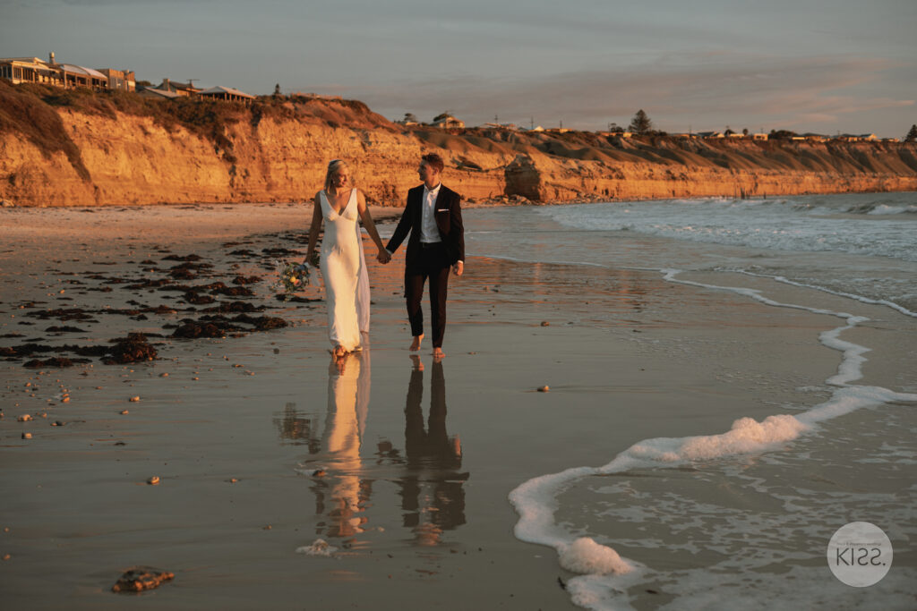 A wedding couple walking along the wet sand beneath coastal cliffs
