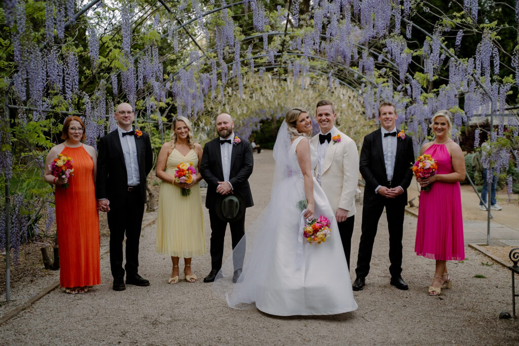 A wedding party standing beneath purple wisteria