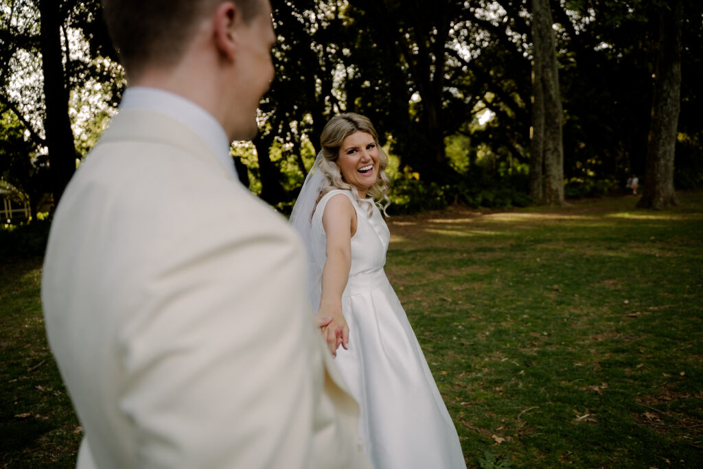 A bride smiling towards her groom beneath garden trees