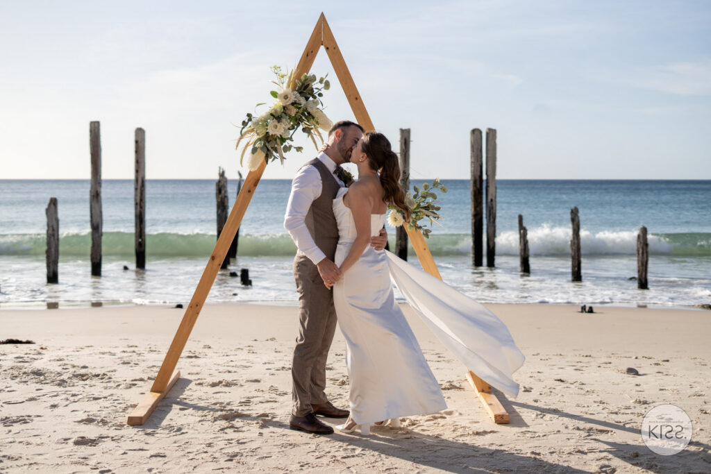 A couple kissing beneath their ceremony arbour at Port Willunga