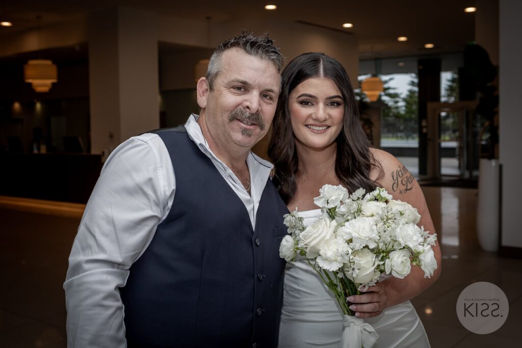 A bride and guest smiling together beside a white wedding bouquet in Glenelg